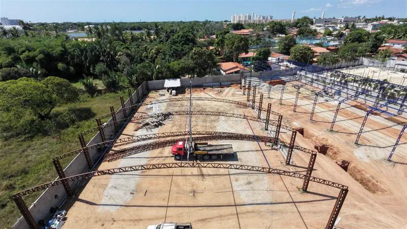 Vista aérea de treliças em arco sendo içadas por caminhão munck durante a montagem da estrutura